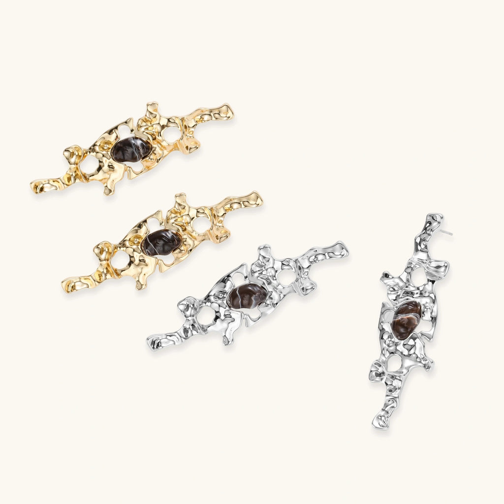 Gold-toned abstract dangle earrings with flowing cutout design and dark resin center accents on white background.